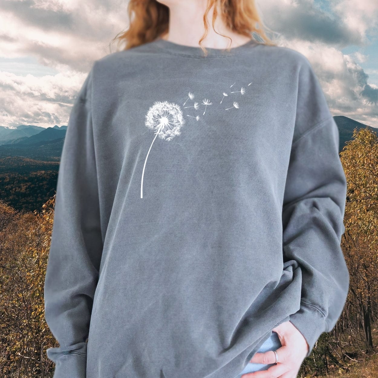 Person wearing Dandelion Comfort Colors Sweatshirt with white dandelion print, showcasing its relaxed fit and pocket style against a natural backdrop.