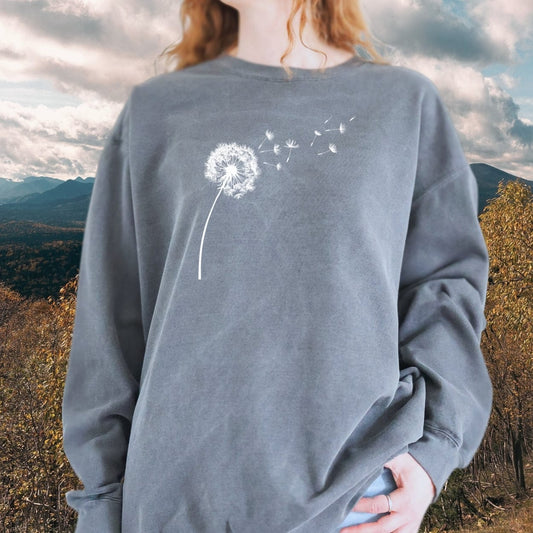 Person wearing Dandelion Comfort Colors Sweatshirt with white dandelion print, showcasing its relaxed fit and pocket style against a natural backdrop.