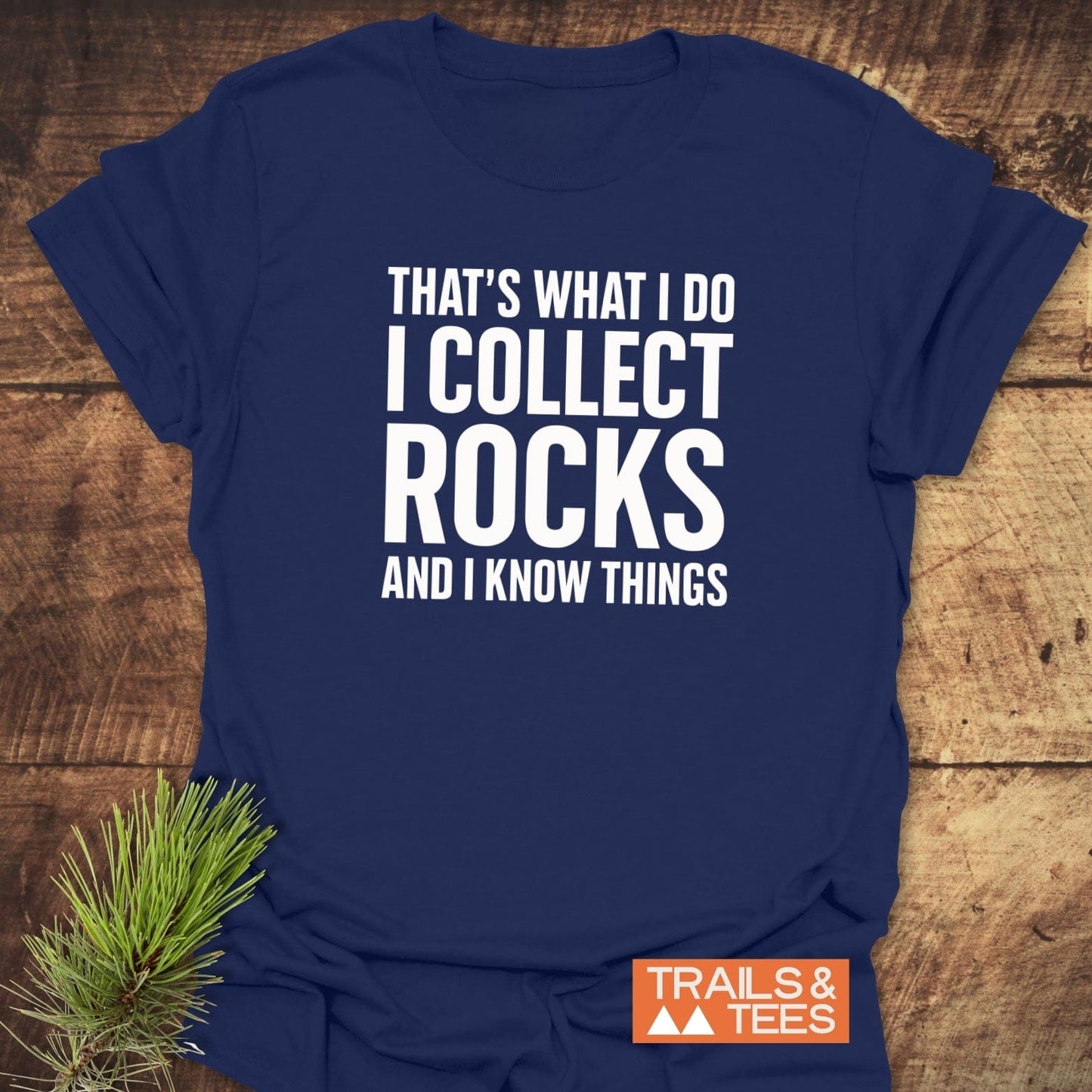 Navy-blue I Collect Rocks T-shirt on wood, featuring bold white text. Includes orange TRAILS & TEES logo. Styled with pine sprig.