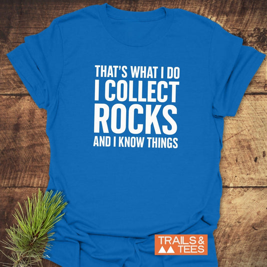 I Collect Rocks T-Shirt featuring bold white slogan, TRAILS & TEES logo, on a wooden surface with pine needles, crafted for outdoor adventures.