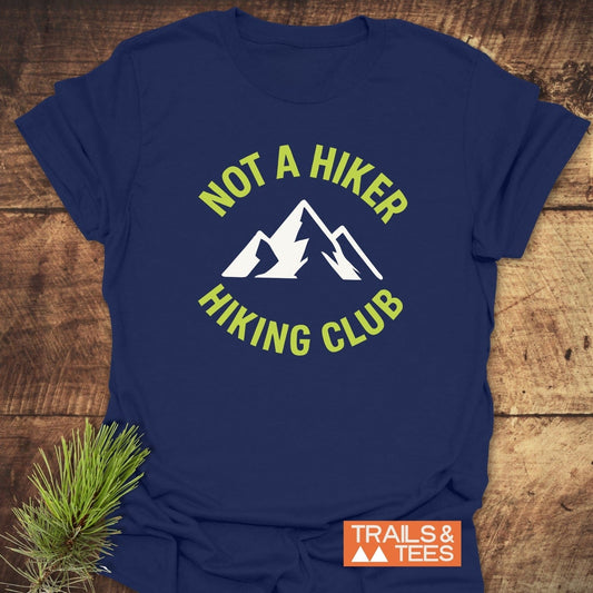Navy Not A Hiker T-Shirt with a white mountain graphic and chartreuse text, laid on wood, featuring a small TRAILS & TEES logo.