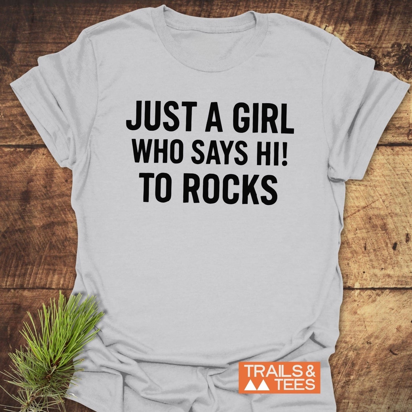 Light-grey Say Hi To Rocks T-shirt with bold black text, Trails & Tees logo, and evergreen sprig, highlighting comfort and outdoor spirit.