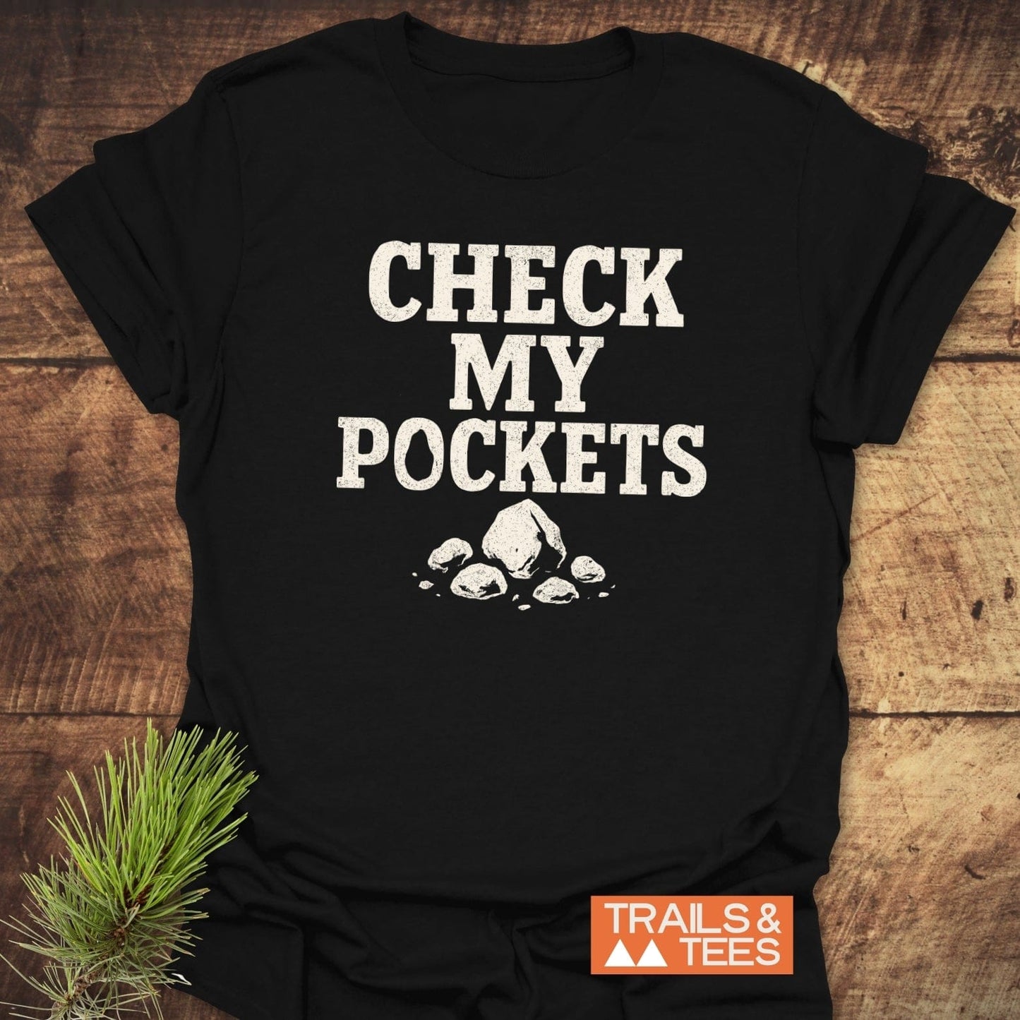 Check My Pockets Rock Lover T-Shirt on wooden surface, featuring distressed white text and rock graphic. Soft ring-spun cotton, durable print, by Trails & Tees.