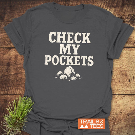 Check My Pockets Rock Lover T-Shirt on wood surface, featuring distressed slogan and rock graphic, styled with pine sprig, showcasing outdoor adventure appeal.