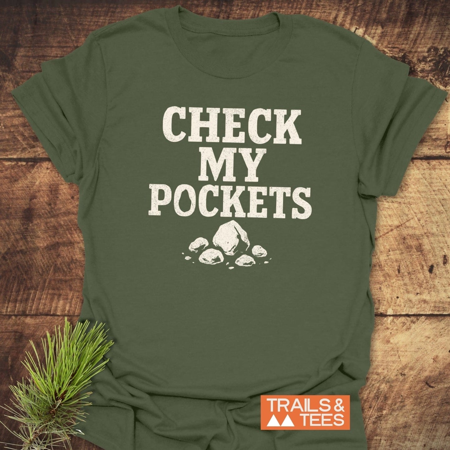 Check My Pockets Rock Lover T-Shirt on rustic surface, featuring distressed white text and rock graphic. Trails & Tees logo visible, pine sprig accentuates outdoorsy theme.