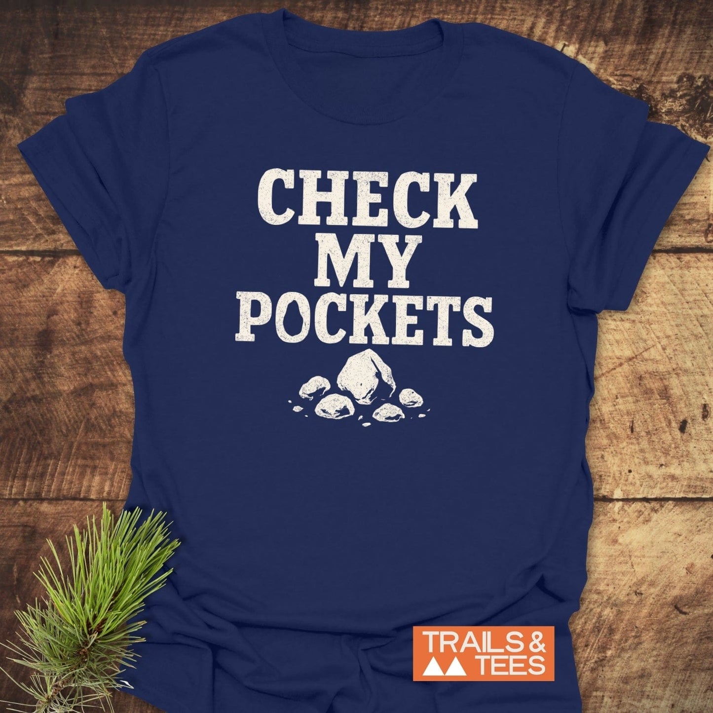 Check My Pockets Rock Lover T-Shirt on wooden surface with bold text and rock graphic, featuring durable, trail-tested comfort from Trails & Tees.