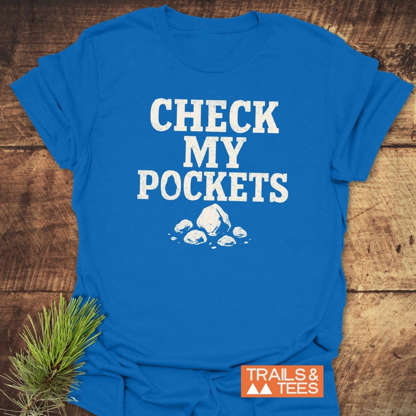 Check My Pockets Rock Lover T-Shirt on wooden surface, features stencil-style text and rock graphic, showcasing durable, soft cotton ideal for outdoor enthusiasts.