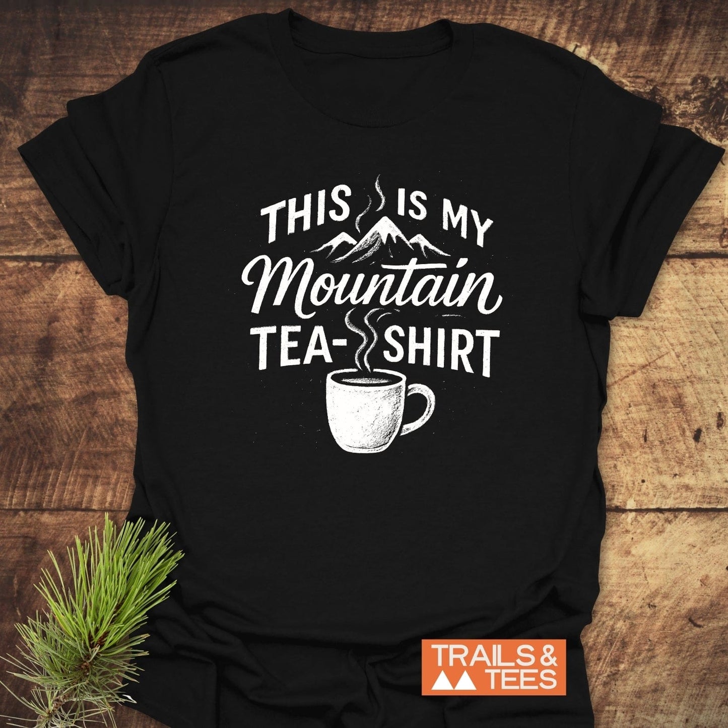 Black Mountain Tea T-Shirt on wood surface, features mountain graphic, slogan, and mug illustration. Includes TRAILS & TEES logo. Crafted for outdoor enthusiasts.