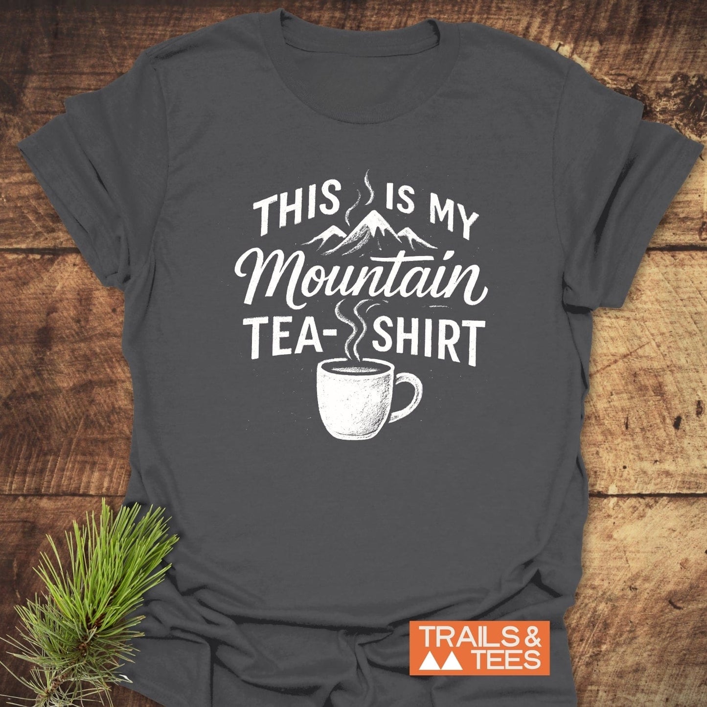 Heather-gray Mountain Tea T-Shirt featuring a mountain graphic and pun slogan. Made from soft cotton, pre-shrunk, with durable prints. Laid on wooden surface.