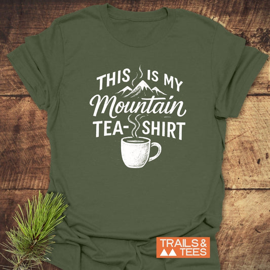 Forest-green Mountain Tea T-Shirt with a white graphic of a tea cup and mountains, laid on wood. Perfect for outdoor adventures from Trails & Tees.