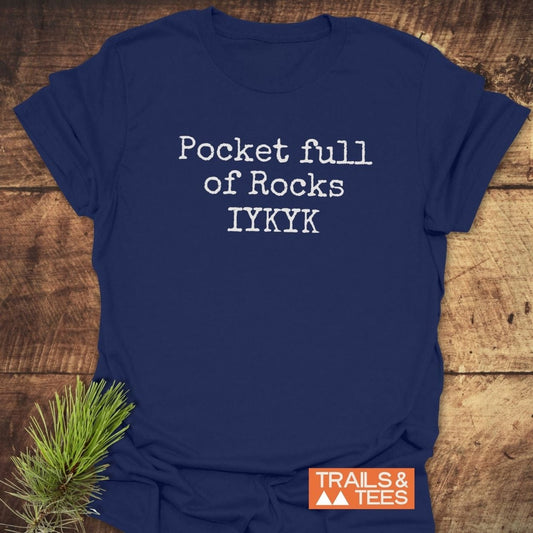 Navy blue tee with Pocket Full Of Rocks IYKYK slogan, styled on wood. Features logo, durable print, and soft cotton for outdoor adventures.