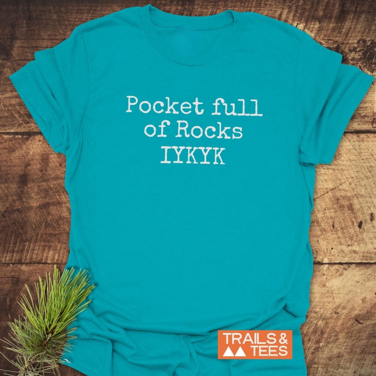Pocket Full Of Rocks IYKYK T-Shirt on wood, with typewriter-style text. Pine branch accent, Trails & Tees logo visible. Perfect for outdoor enthusiasts.