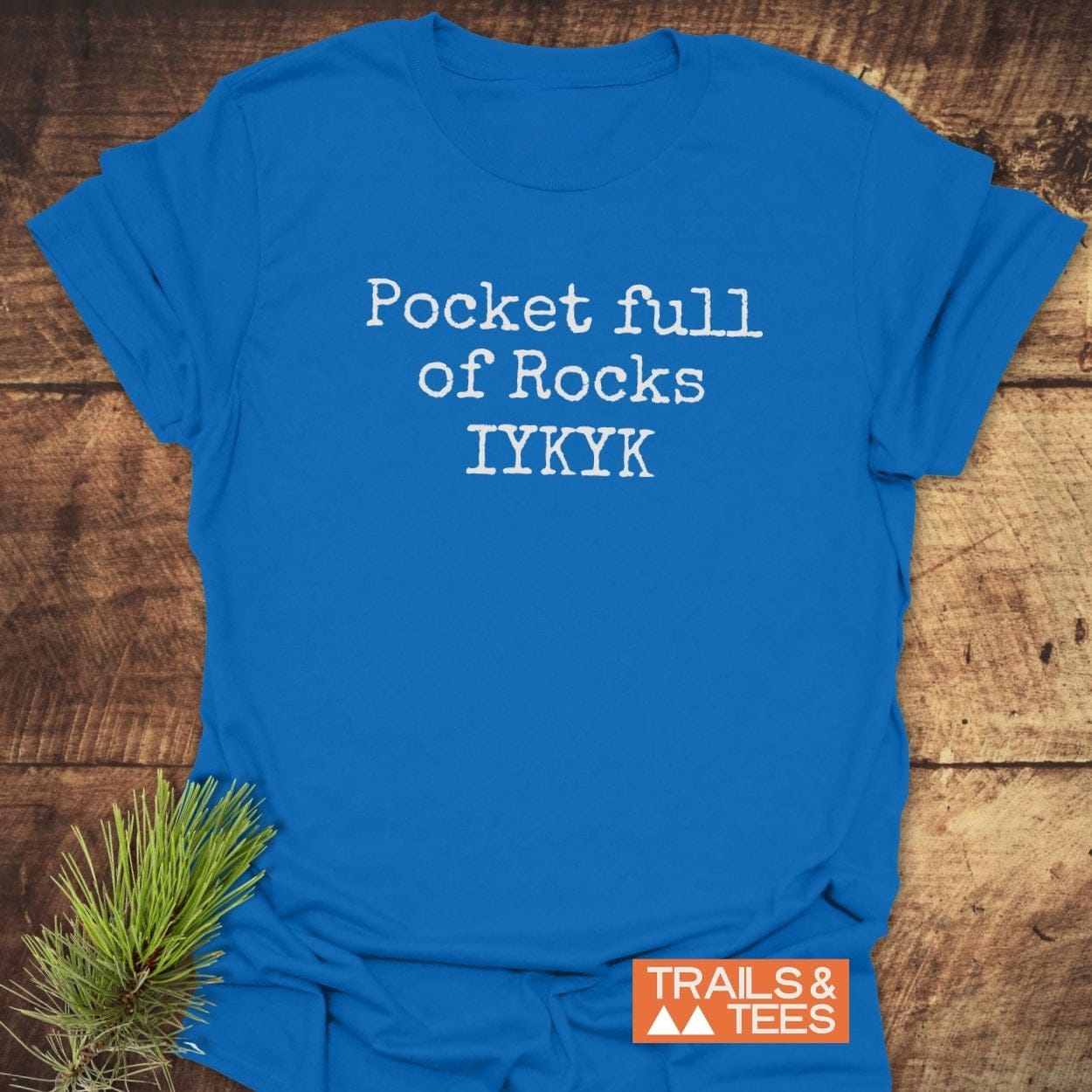Bright blue Pocket Full Of Rocks IYKYK T-shirt on rustic wood, featuring durable white typewriter-style text. Includes TRAILS & TEES. logo, emphasizing comfort and outdoor-ready design.