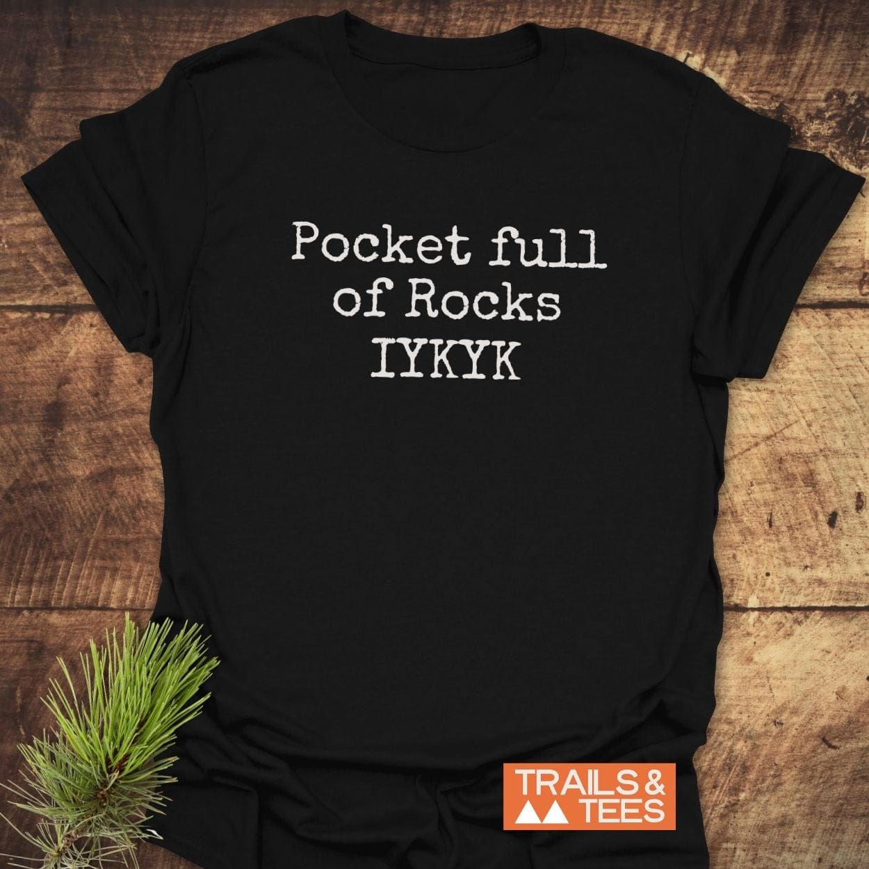 Black t-shirt with Pocket Full Of Rocks IYKYK in white typewriter-style text. Features soft, durable cotton for outdoor adventures. Logo TRAILS & TEES. at bottom right.