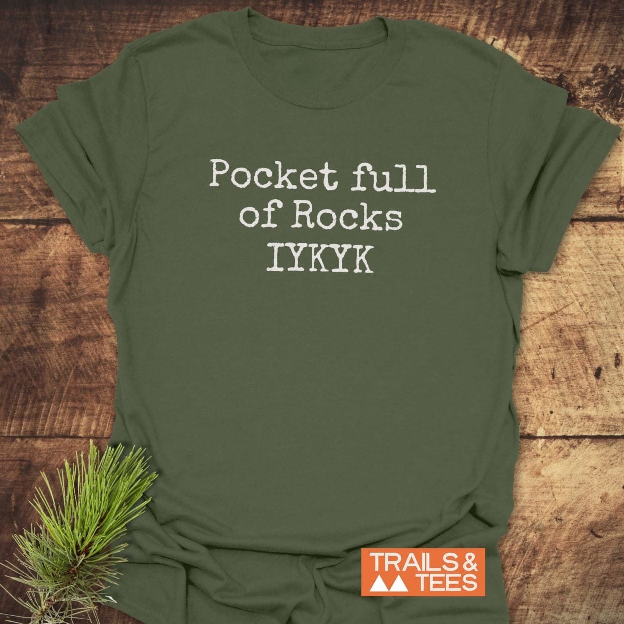 Olive-green T-shirt with Pocket Full Of Rocks IYKYK in white font, featuring a small TRAILS & TEES logo, displayed on a wooden surface.