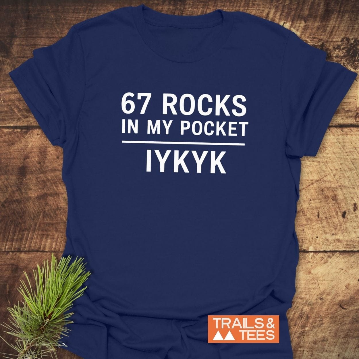 Navy-blue 67 Rocks In My Pocket T-shirt with white print, laid on wood. Features durable print, no side seams, and TRAILS & TEES logo.