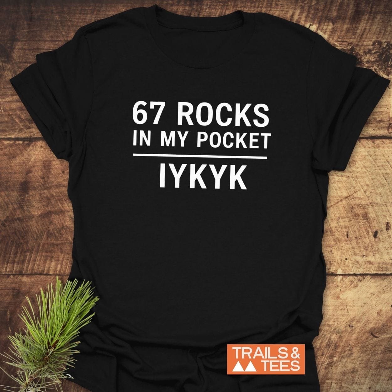 Black t-shirt with “67 ROCKS IN MY POCKET” in bold white text, featuring Trails & Tees logo and pine sprig; crafted for outdoor adventures.