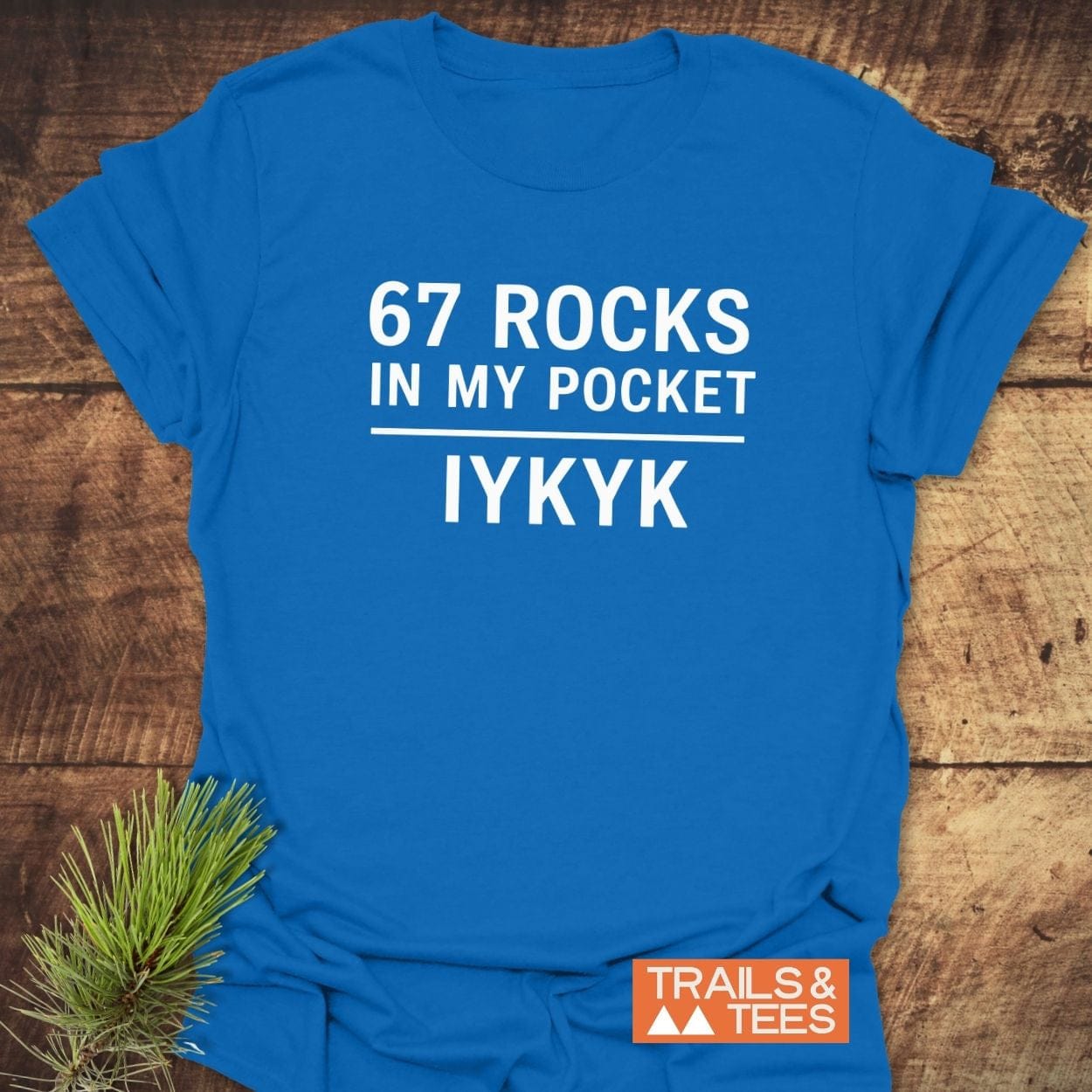 67 Rocks In My Pocket T-Shirt on rustic surface, featuring bold white slogan, Trails & Tees logo, and pine sprig, emphasizing outdoor adventure and comfort.