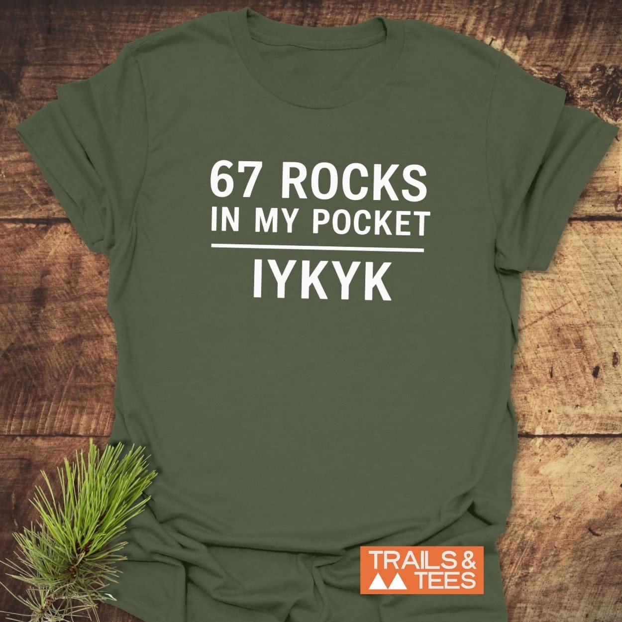 67 Rocks In My Pocket T-Shirt in olive-green, features white lettering and Trails & Tees logo, styled with pine needles, crafted for outdoor enthusiasts.