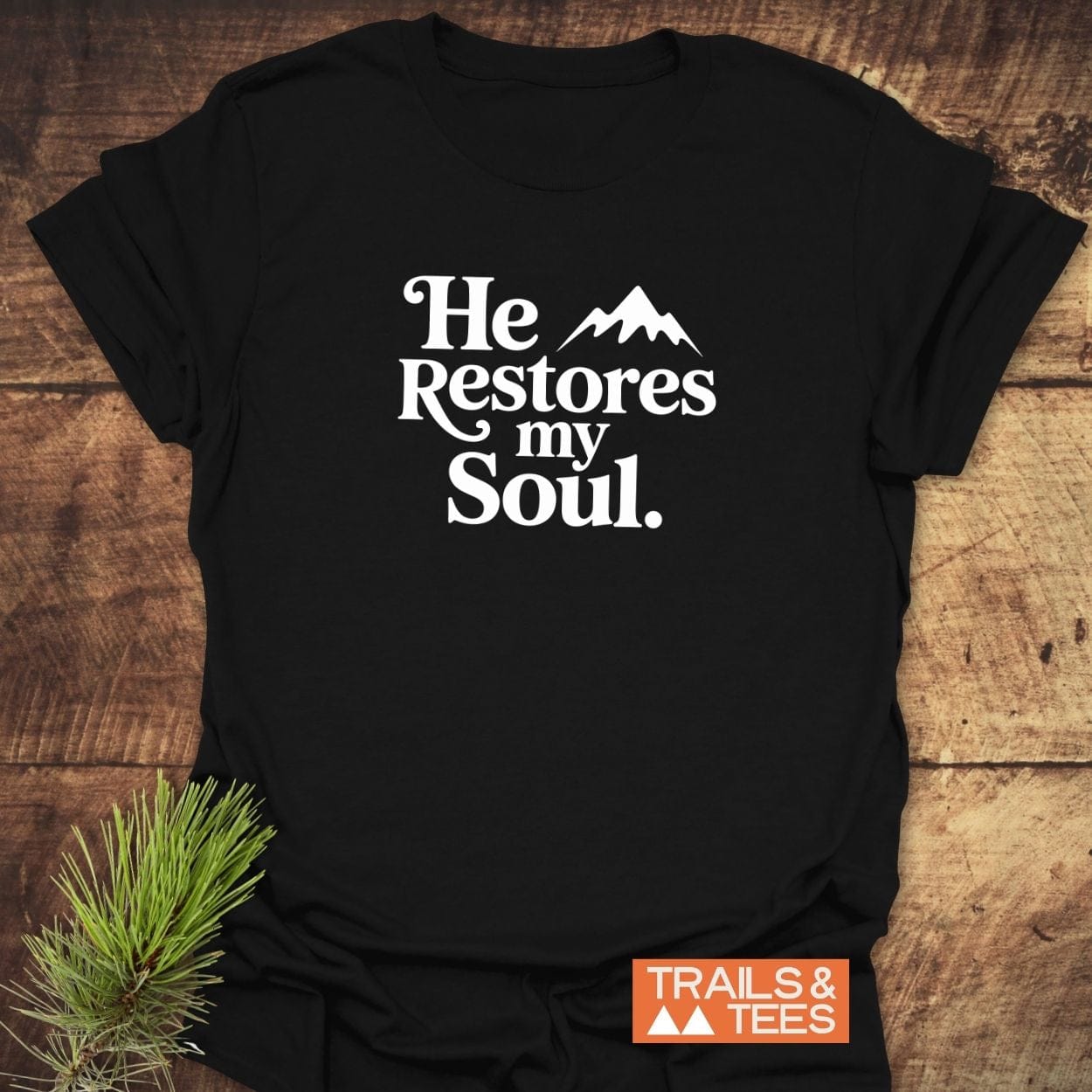 Black T-shirt with He Restores my Soul text and mountain graphic, showcasing outdoor adventure appeal. Pine needles styled beside for a natural touch.