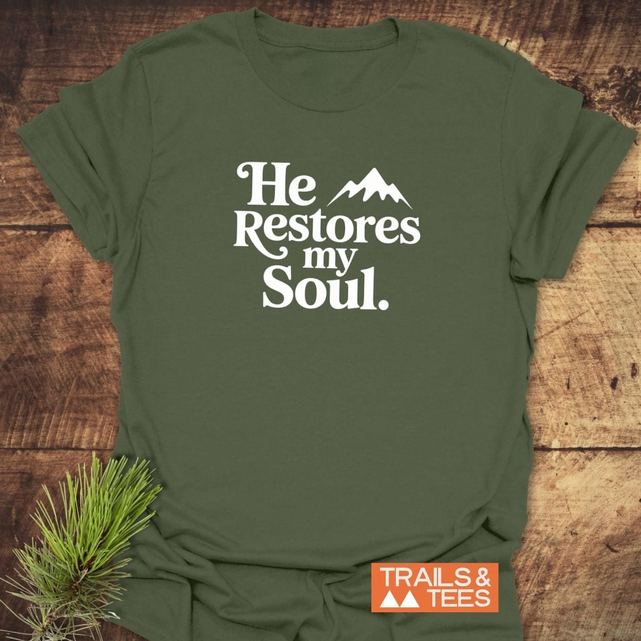 He Restores My Soul T-Shirt in olive-green, featuring a white graphic and mountain silhouette, laid flat with Trails & Tees logo and pine sprig accent.
