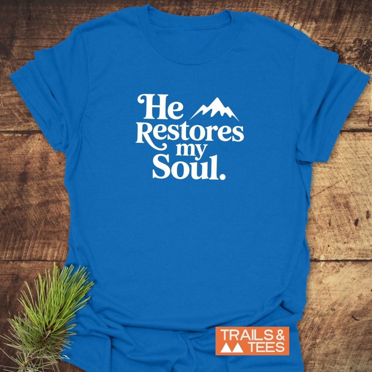 “He Restores My Soul” T-shirt featuring a mountain design, crafted from soft cotton, laid on wood with an orange Trails & Tees logo.