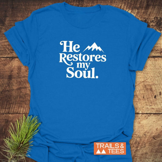 “He Restores My Soul” T-shirt featuring a mountain design, crafted from soft cotton, laid on wood with an orange Trails & Tees logo.