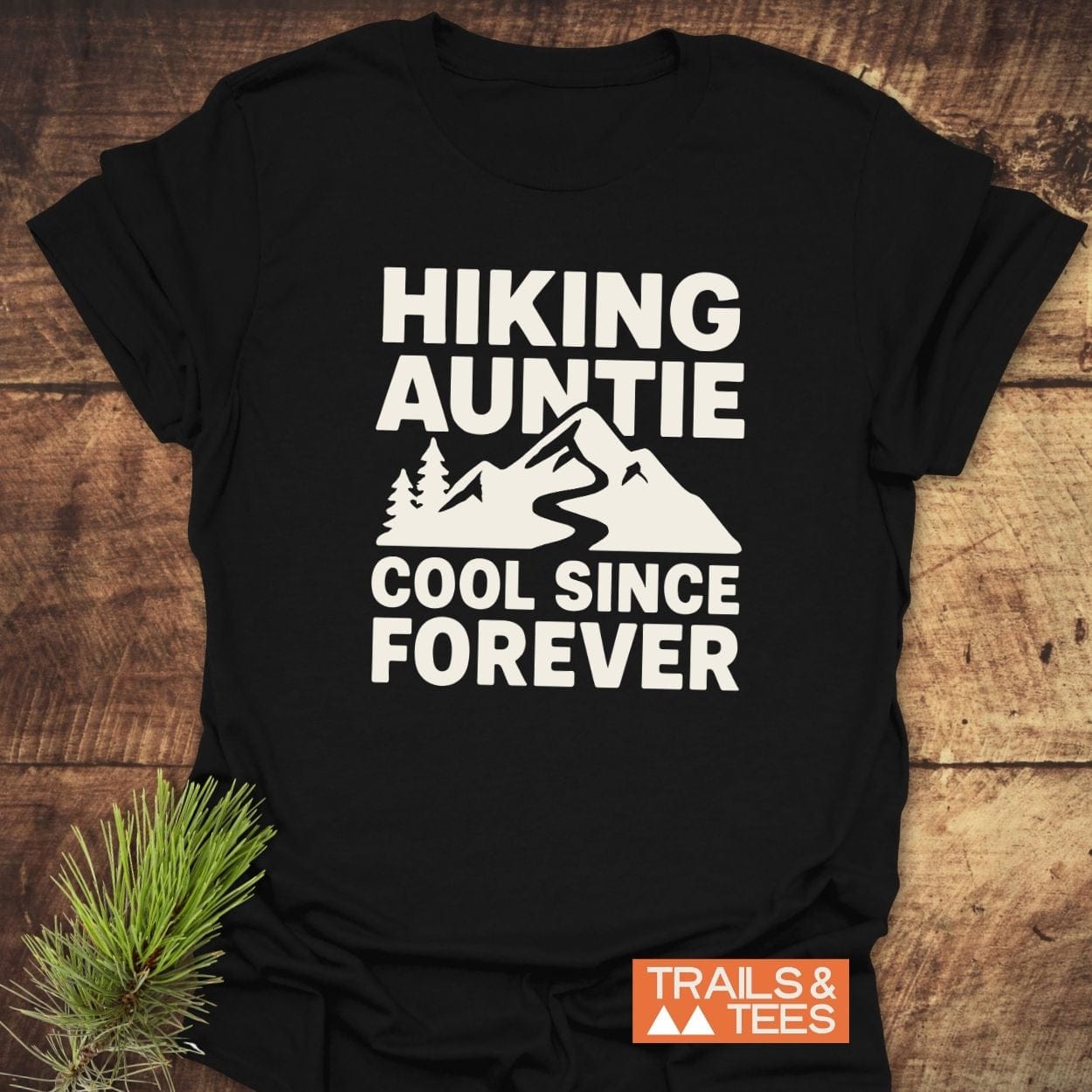 Hiking Auntie T-Shirt with bold graphic, featuring mountains and trees, on soft black cotton. Includes “TRAILS & TEES” logo. Pine needles beside shirt.