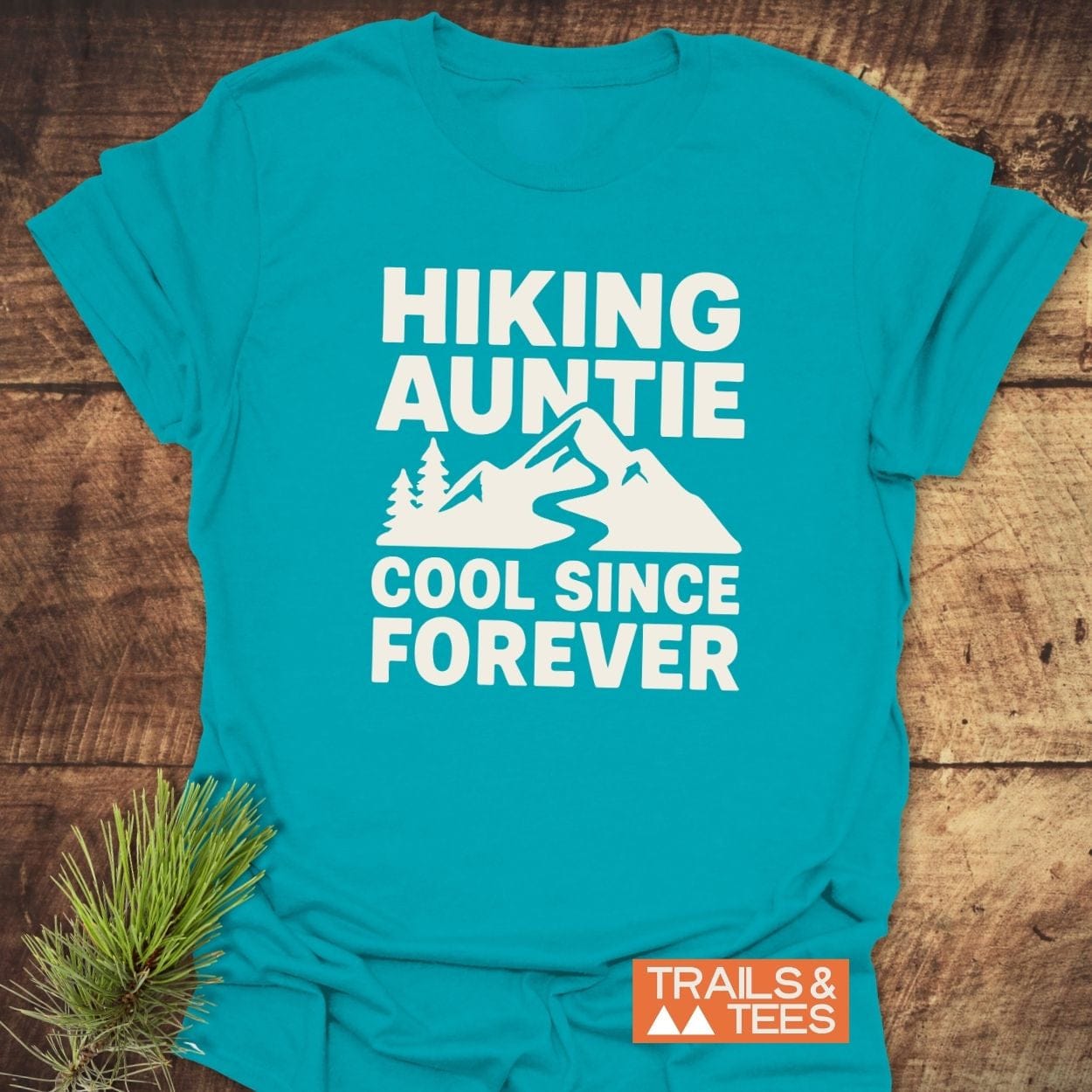 Hiking Auntie T-Shirt featuring a white mountain graphic and text on a teal-blue background, showcasing a durable, adventure-ready design by Trails & Tees.