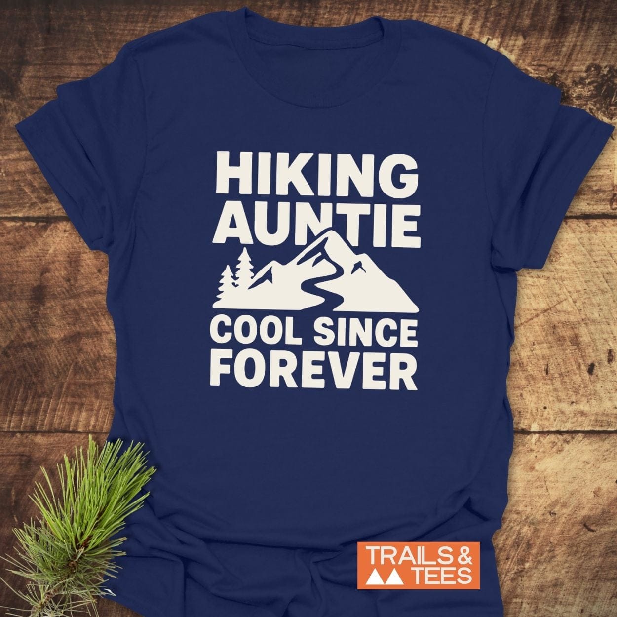 Hiking Auntie T-Shirt featuring a mountain peak graphic and text, designed for comfort and durability, laid on a wood background with pine sprig.