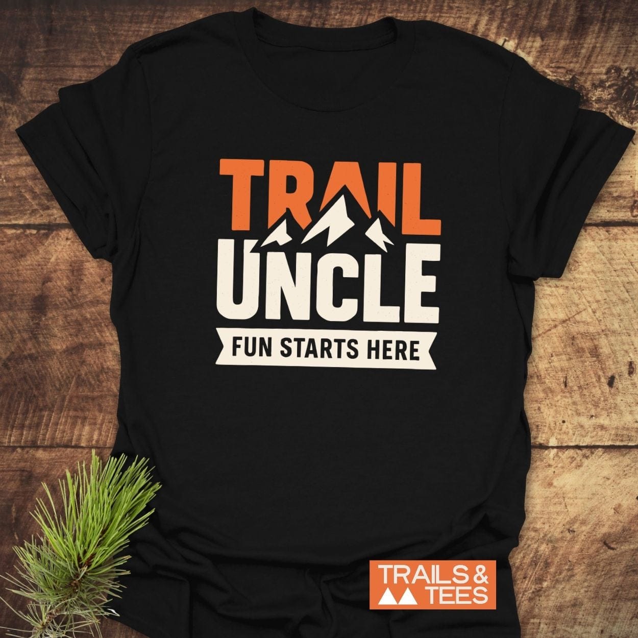 Trail Uncle Funny T-Shirt with mountain silhouette and bold graphic, displayed on wood with pine sprig. Crafted from soft cotton for outdoor enthusiasts.