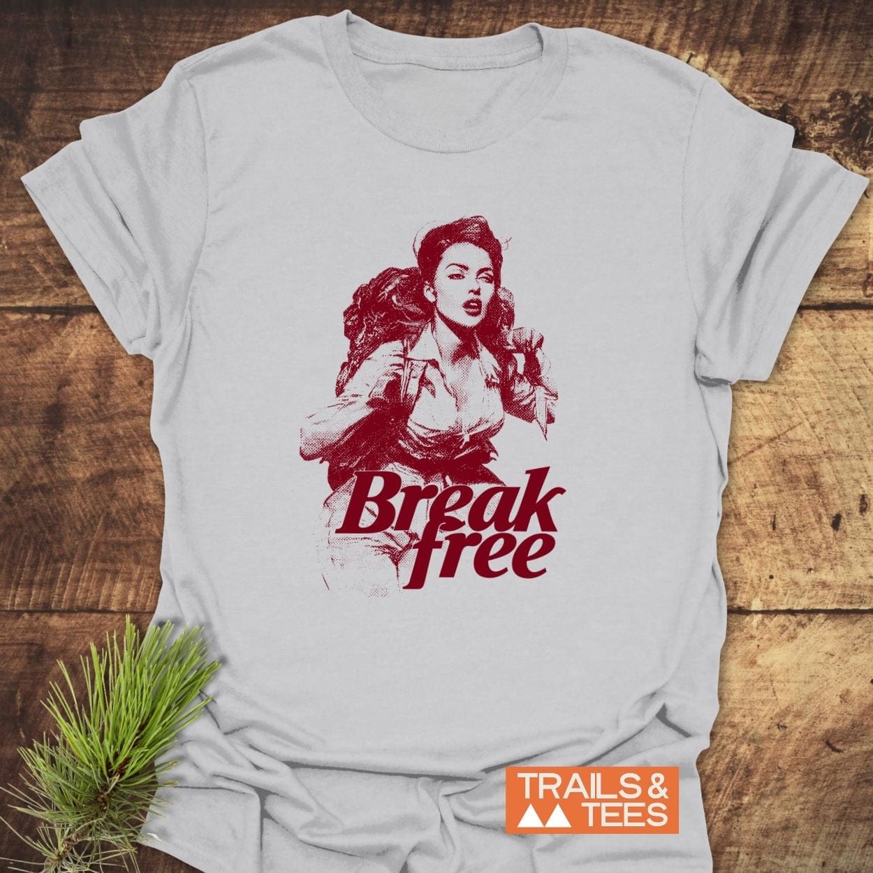 Break Free Hiking T-Shirt featuring a vintage-style red halftone hiker print on light-gray cotton, laid on wood with a pine sprig, by TRAILS & TEES.
