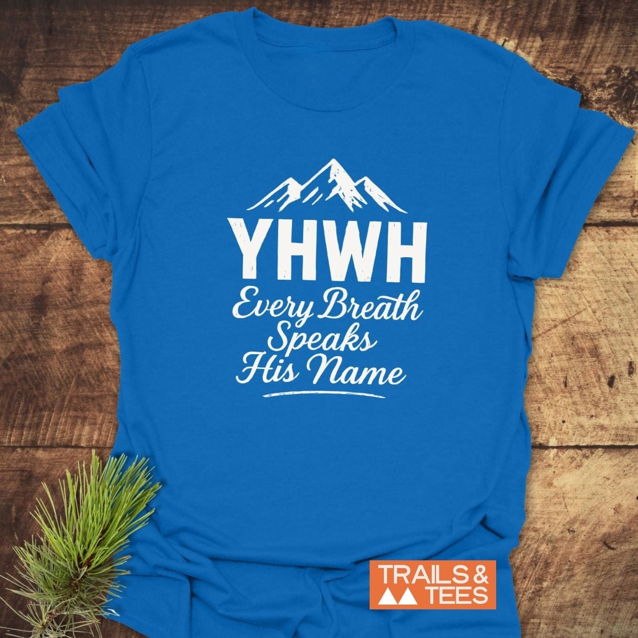 YHWH Christian Hiking T-Shirt with mountain graphic and “Every Breath Speaks His Name” text, laid on wood. Includes “TRAILS & TEES” logo and pine needles.