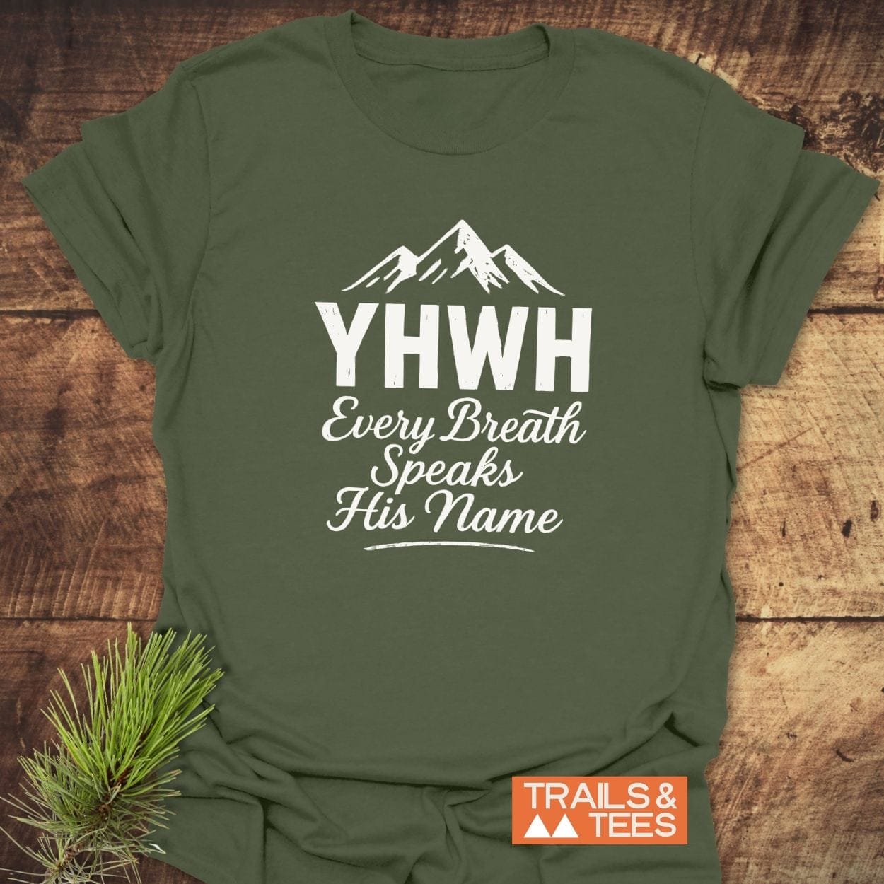 YHWH Christian Hiking T-Shirt with mountain graphic, Every Breath Speaks His Name text, on wooden surface beside pine sprig, featuring durable, trail-tested design.