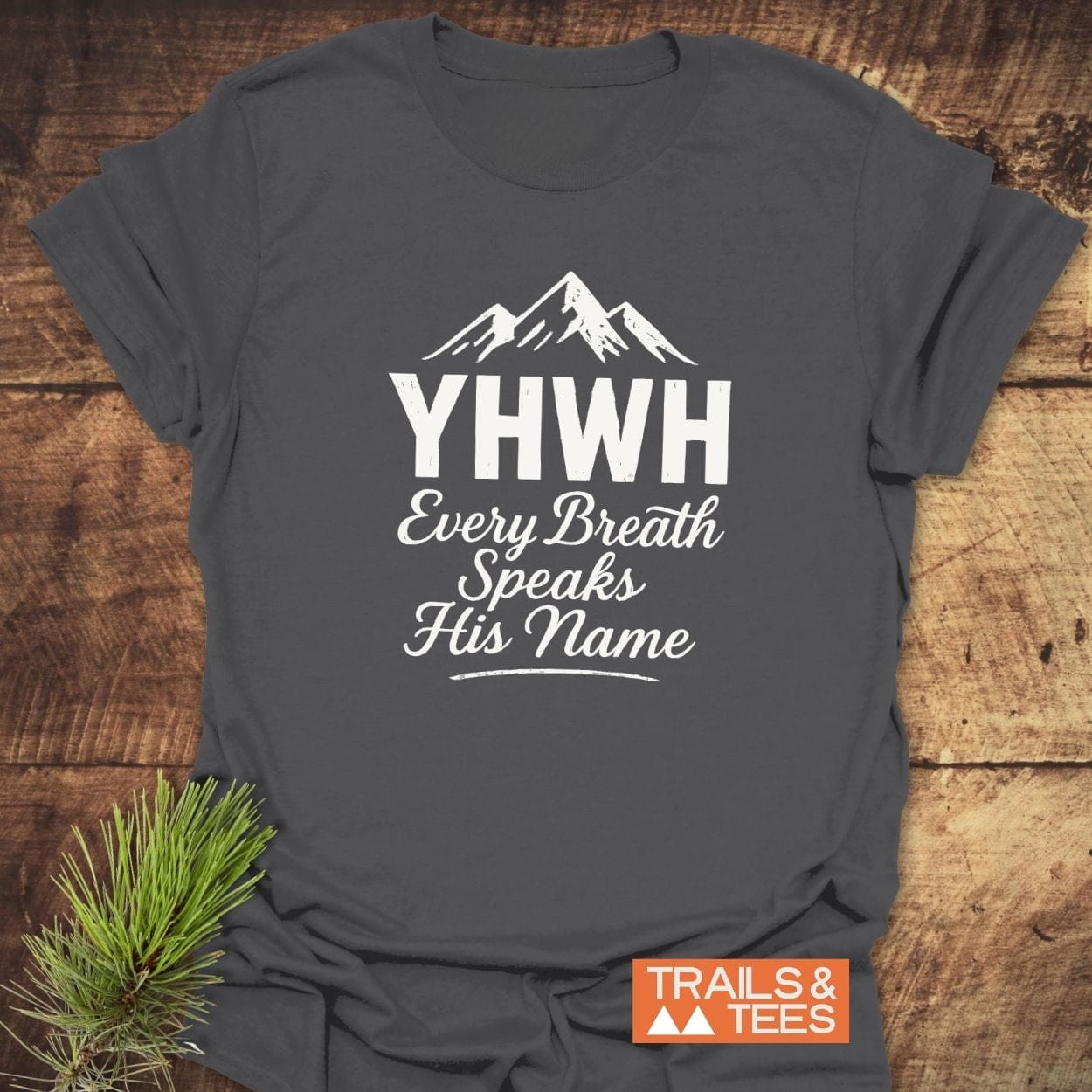 Charcoal T-shirt with YHWH and mountain graphic; Every Breath Speaks His Name. Styled with pine sprig. Made from soft, lightweight cotton for outdoor enthusiasts.