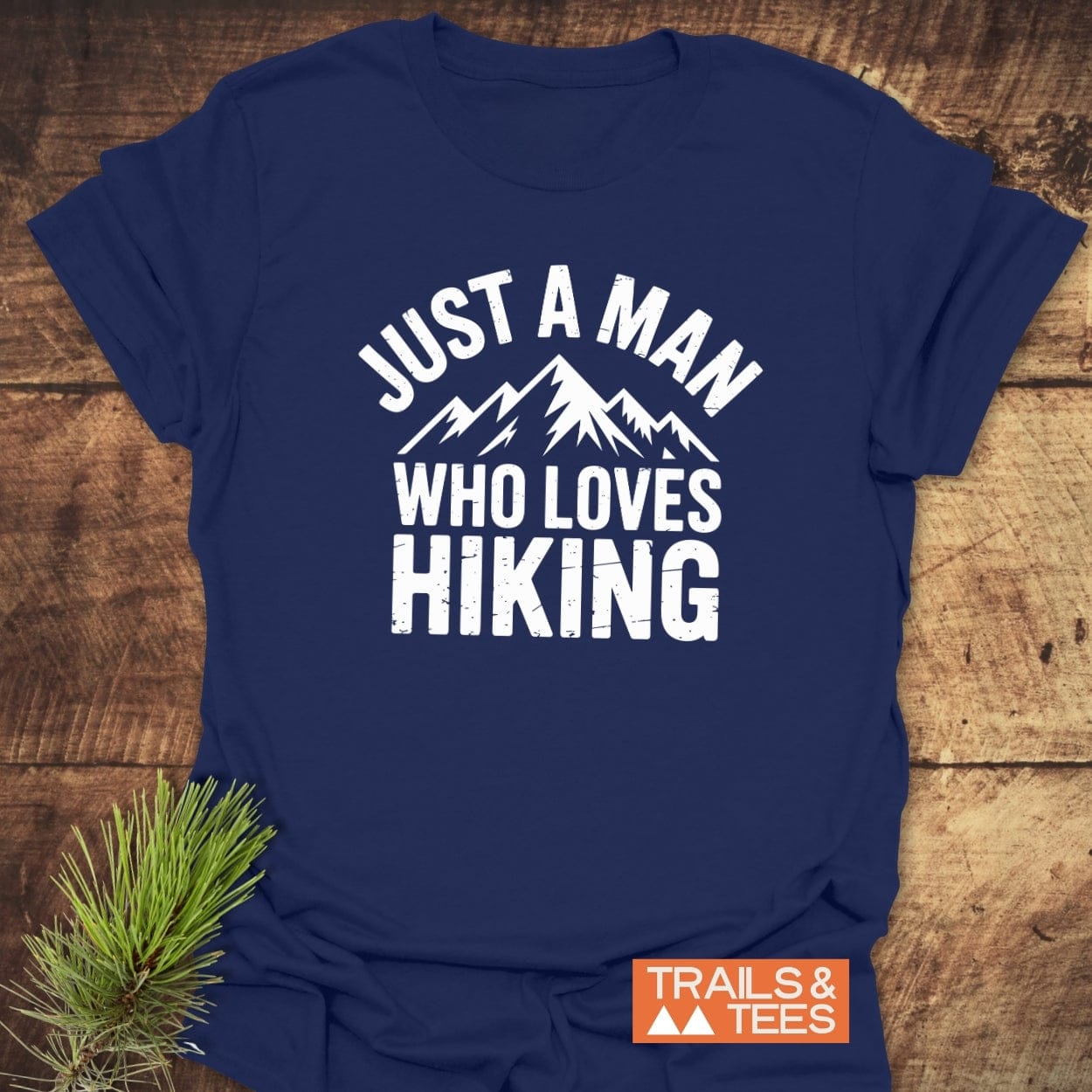 Navy-blue Just A Man Who Loves Hiking T-shirt, featuring a mountain silhouette, laid flat with TRAILS & TEES logo and pine sprig accent.