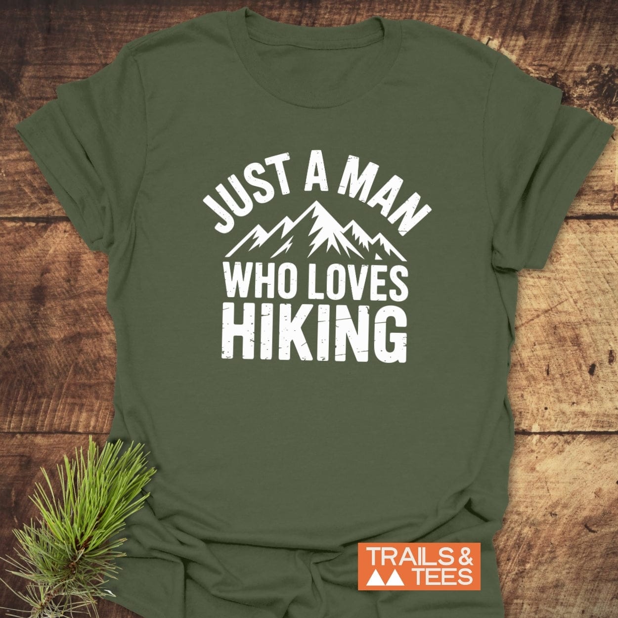 Just A Man Who Loves Hiking T-Shirt flat lay featuring mountain graphic, pine sprigs; crafted from soft ring-spun cotton for comfort and durability.