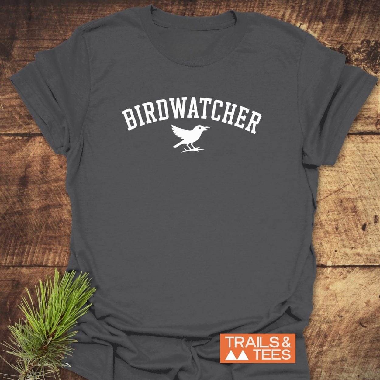 Birdwatcher Varsity T-Shirt on wooden surface, featuring BIRDWATCHER text and bird silhouette. Highlighting comfort, durability, and eco-friendly design from Trails & Tees.