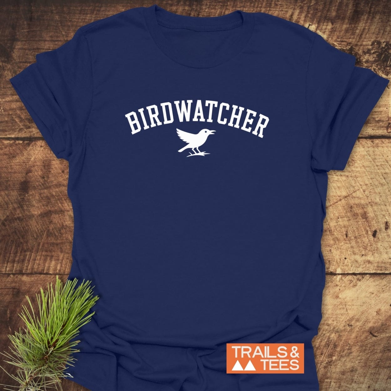 Birdwatcher Varsity T-Shirt featuring white block letters and bird silhouette on navy-blue, styled with a pine sprig. Comfortable, pre-shrunk, ring-spun cotton for outdoor adventures.