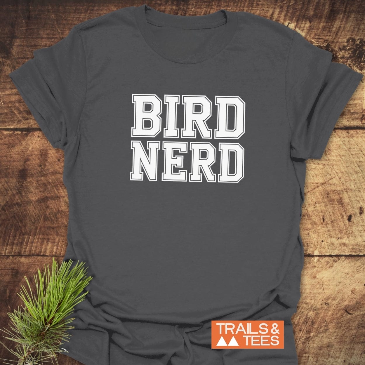 Bird Nerd Varsity T-Shirt on rustic wood, featuring bold white varsity-style lettering and a pine sprig accent, embodying outdoor adventure and comfort.