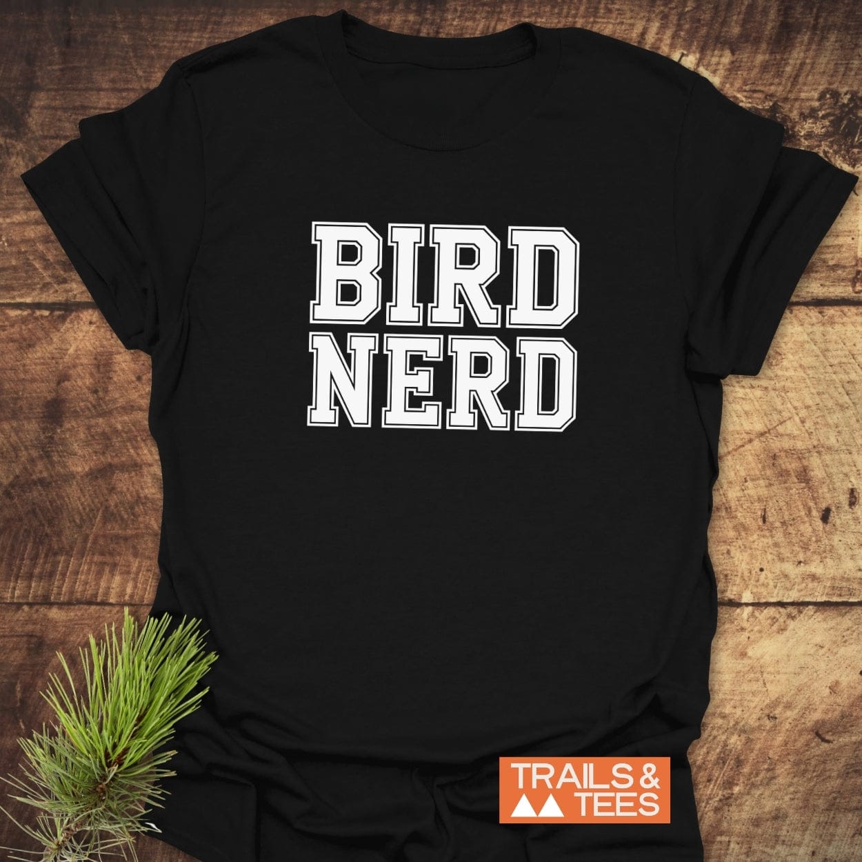 Bird Nerd Varsity T-Shirt, black with bold white letters, lies flat with a small Trails & Tees logo and pine greenery accentuating its adventurous spirit.