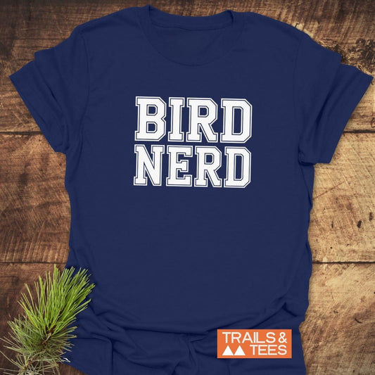 Bird Nerd Varsity T-Shirt on wooden surface, featuring white block slogan and “TRAILS & TEES” logo. Accompanied by pine sprig, highlighting outdoor adventure theme.