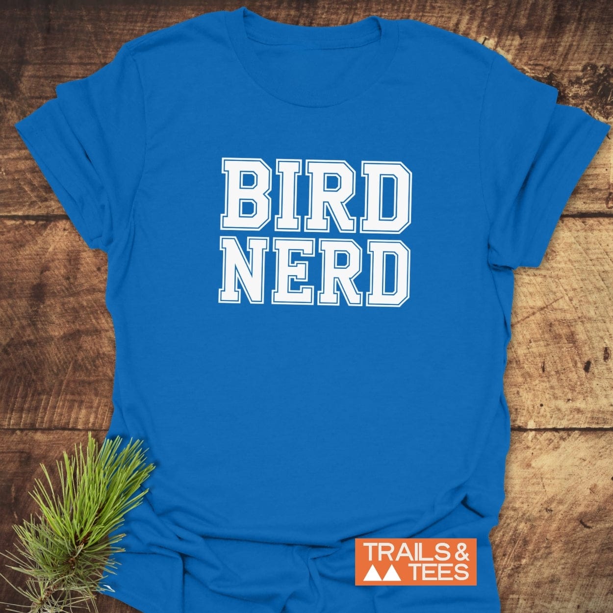 Bird Nerd Varsity T-Shirt on wooden surface, featuring white block letters, orange Trails & Tees logo, and pine sprig, showcasing soft, durable outdoor-friendly design.