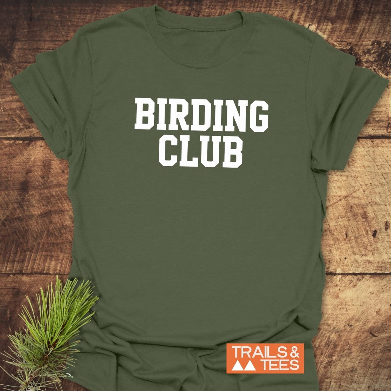 Birding Club T-Shirt on rustic wood, featuring bold white collegiate text. Includes pine bough accent and Trails & Tees logo, highlighting outdoor adventure wear.