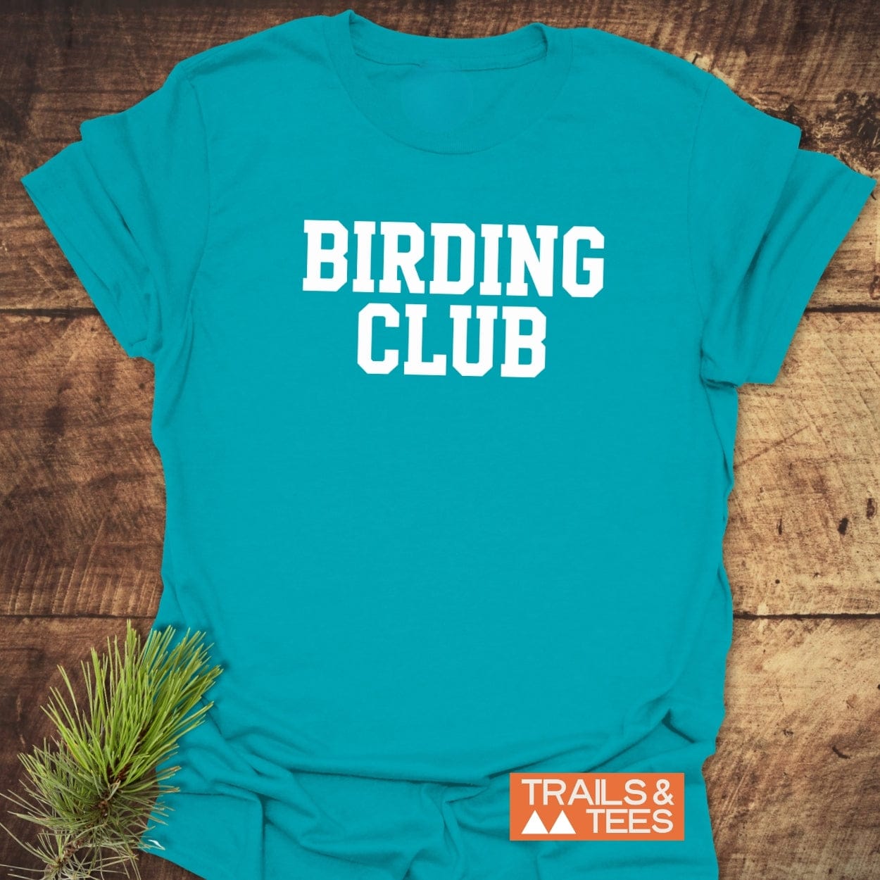 Birding Club T-Shirt in teal on wood, featuring bold white lettering and an orange Trails & Tees logo, accented by an evergreen sprig.