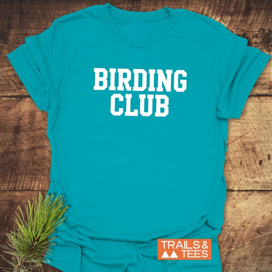 Birding Club T-Shirt in teal on wood, featuring bold white lettering and an orange Trails & Tees logo, accented by an evergreen sprig.