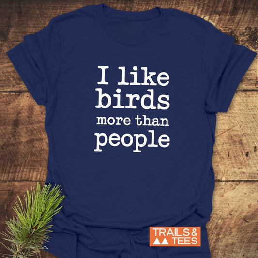 Navy-blue I Like Birds More Than People T-shirt on wooden surface, featuring durable print and TRAILS & TEES tag, accented by a pine sprig.