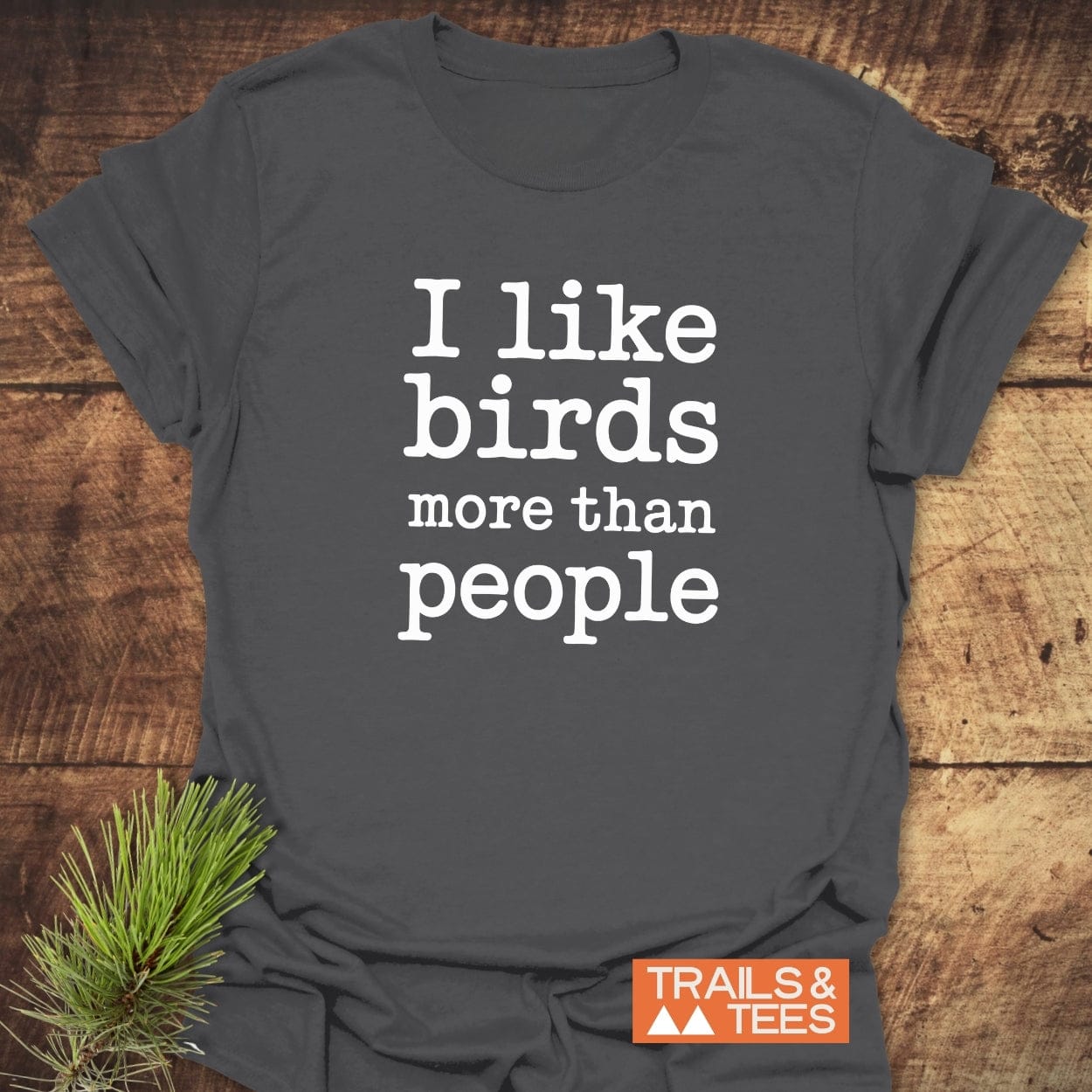 Dark-gray I Like Birds More Than People T-shirt on wood, featuring large white text. Includes Trails & Tees logo and pine sprig for outdoorsy appeal.