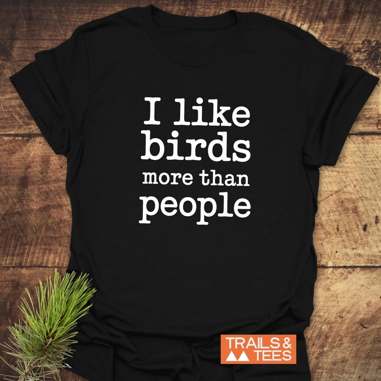 I Like Birds More Than People T-Shirt on wood, featuring bold white slogan and orange TRAILS & TEES logo. Styled with pine sprig for outdoorsy appeal.
