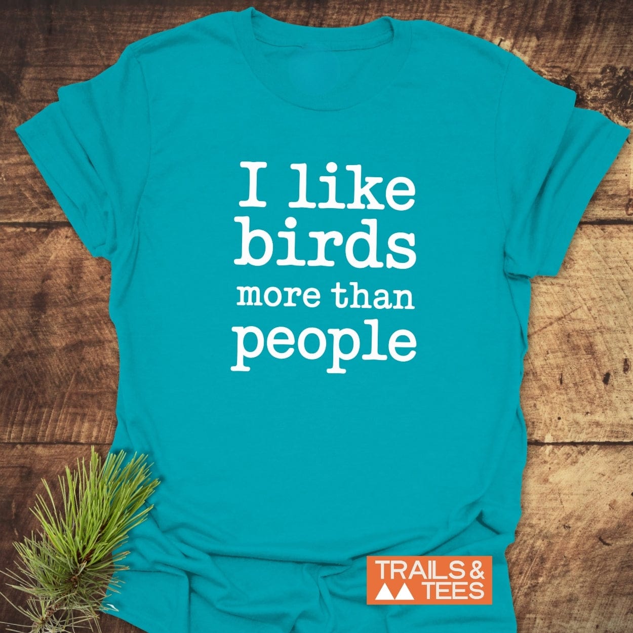 Teal-blue t-shirt with I Like Birds More Than People in white letters, featuring Trails & Tees logo; pine sprig beside for an outdoor vibe.